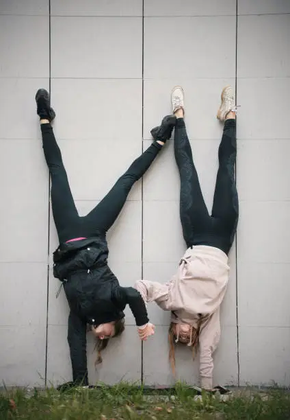 Two girls acrobats holding hands performing a handstand against the background of the wall. Wide shot