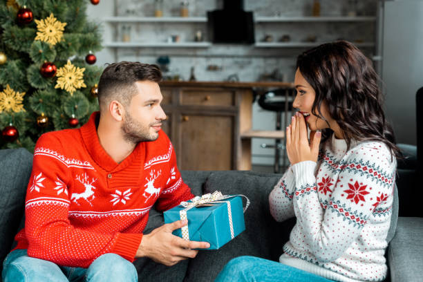 istock handsome young man gifting christmas present to surprised girlfriend gesturing by hands on sofa at home 1048803388