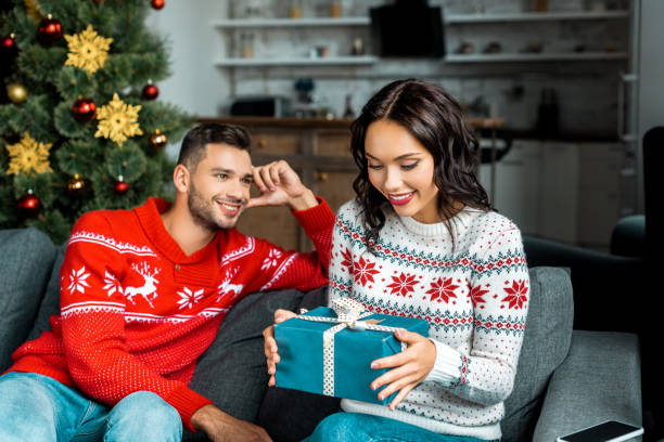 istock attractive young woman untying christmas present while her boyfriend sitting on sofa behind at home 1048803424