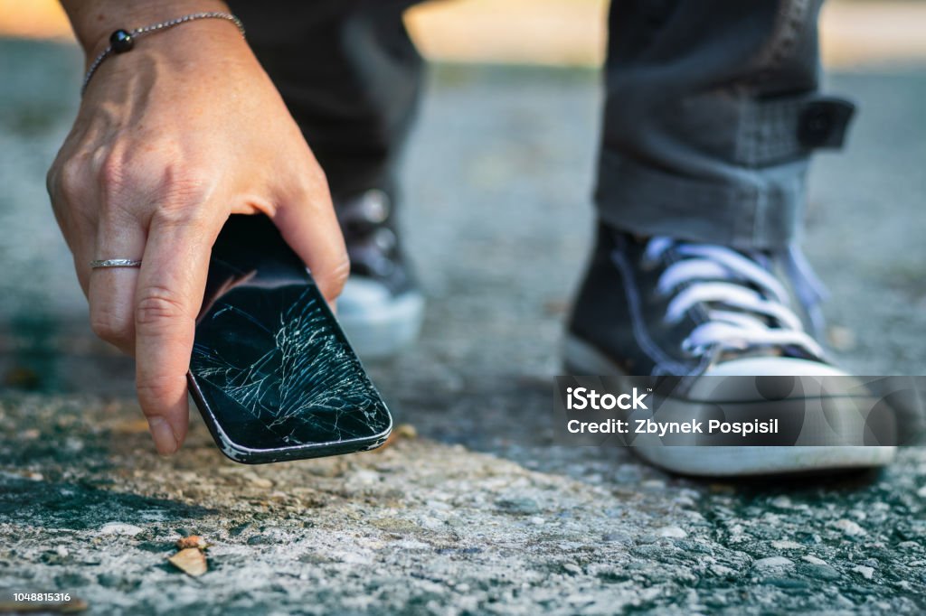 Woman picking up broken smartphone from the ground. Damaged mobile phone with cracked touch screen. Telephone Stock Photo