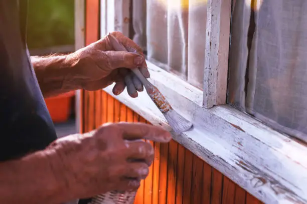 Photo of Senior man painting wooden windows using paintbrush. Repairing exterior of old house.