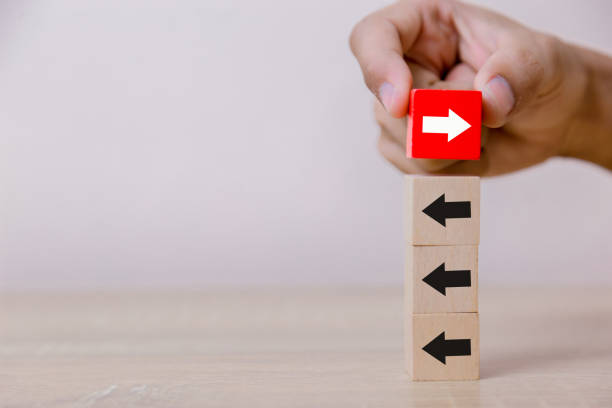 Hand putting wood cube block on top vertical red wooden blocks with white arrows facing opposite to the black arrows. With different concepts to other people. Going for success. Hand putting wood cube block on top vertical red wooden blocks with white arrows facing opposite to the black arrows. With different concepts to other people. Going for success. changing focus stock pictures, royalty-free photos & images