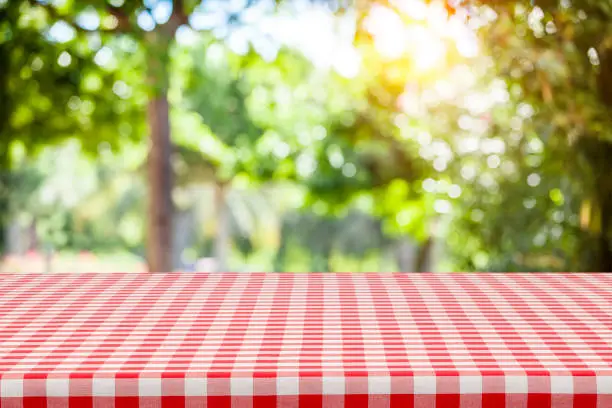 Empty table covered with red and white checkered tablecloth with defocused lush foliage at background. Ideal for product display on top of the table. Predominant color are green and red. DSRL studio photo taken with Canon EOS 5D Mk II and Canon EF 100mm f/2.8L Macro IS USM.
