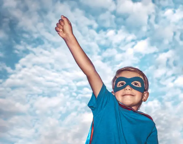 Portrait of a little boy wearing costume of superhero with  raised up one hand over sky background, is preparing to save the world, kid plays superman