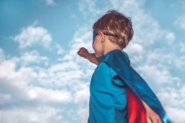 Little boy wearing costume of a superhero with  raised up one hand over sky background, preparing to save the world, kid plays in superman