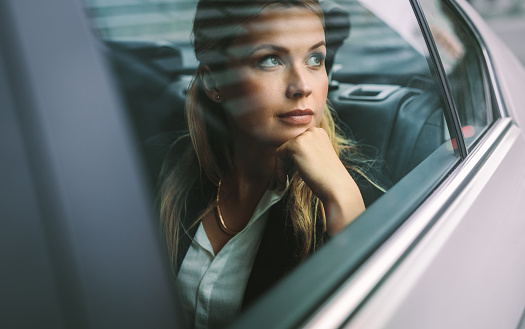 Beautiful young businesswoman sitting on back seat of a car and looking outside the window. Female business executive travelling by a cab.
