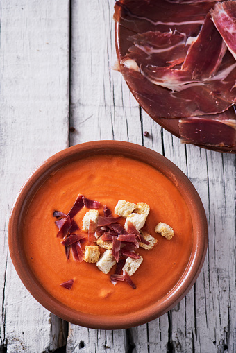 high angle view of an earthenware bowl with spanish salmorejo cordobes or porra antequerana, a cold tomato soup, topped with serrano ham and croutons, on a white rustic table