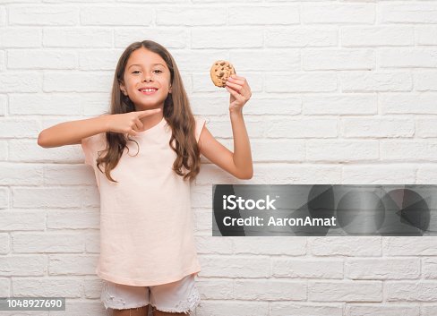 istock Young hispanic kid over white brick wall eating chocolate chips cooky very happy pointing with hand and finger 1048927690