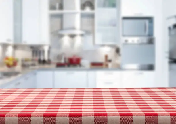 Empty kitchen countertop covered with red and white checkered tablecloth with blurred modern kitchen at background. Predominant colors are white and red. Very suitable for product montage.