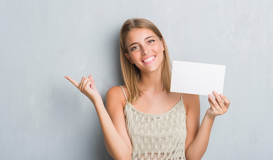 Beautiful young woman over grunge grey wall holding blank card very happy pointing with hand and finger to the side