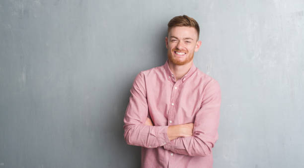 młody rudowłosy mężczyzna na szarej ścianie grunge ubrany w różową koszulę szczęśliwą twarz uśmiechniętą ze skrzyżowanymi ramionami patrzącą na kamerę. pozytywna osoba. - arms crossed zdjęcia i obrazy z banku zdjęć