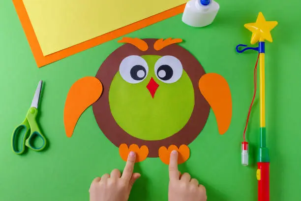 making of diy lantern for sankt martin, traditional german feast, childs hands and owl of color paper, glue, scissors, lanterns wand on green background