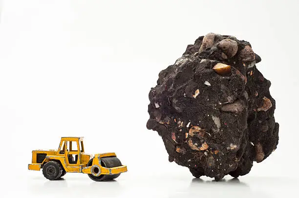 Miniature toy car road roller (roller-compactor, roller) next to a relatively huge piece of asphalt. Concept for road construction and foundation work. Type of road roller: ride-on articulating-swivel.