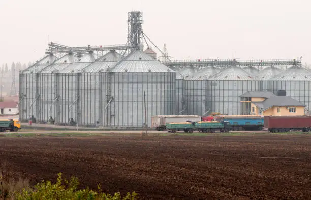 Plowed field in front of grain elevator. Grain trucks near the agrocomplex.