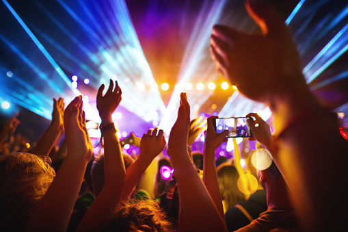 Rear view of excited crowd enjoying a DJ performance at a festival. There are many raised hands, some of the holding cell phones and taping the show.
People in foreground are released.