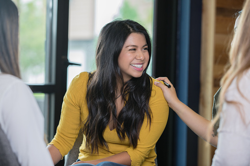 Beautiful Hispanic young adult woman is smiling during a support group therapy meeting. Unrecognizable woman is placing her hand on shoulder to offer support.