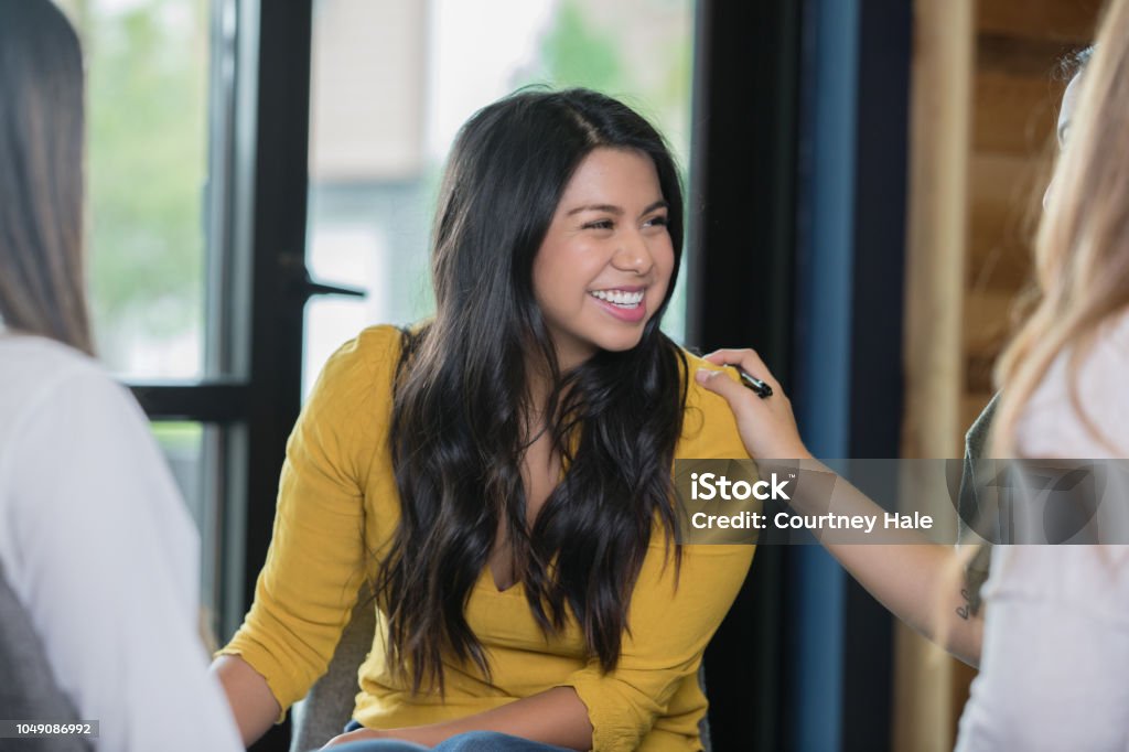 Hispanic woman smiling during support group therapy meeting Beautiful Hispanic young adult woman is smiling during a support group therapy meeting. Unrecognizable woman is placing her hand on shoulder to offer support. Group Therapy Stock Photo