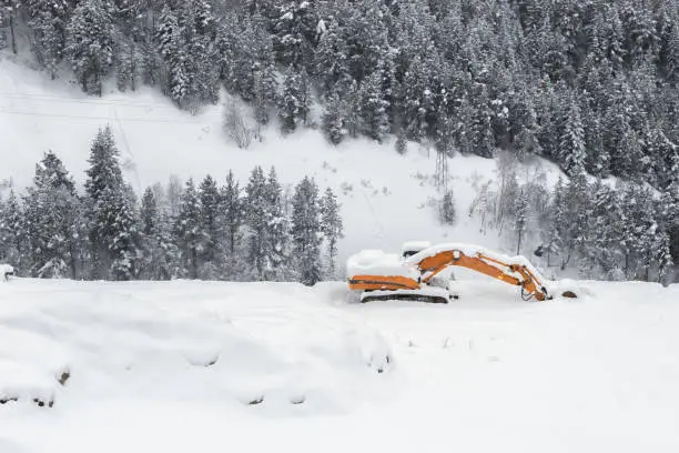 The excavator covered with snow against the background of snow-covered fir-trees. Heavy snowfall, big snowdrifts of snow in the winter
