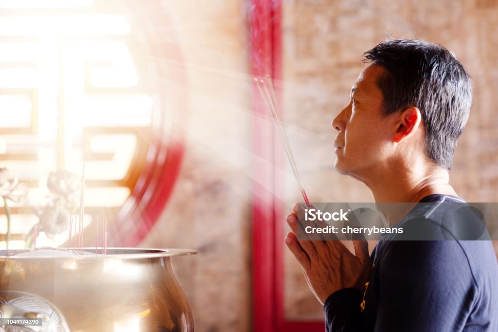 Faith and religious. Man hands holding joss sticks blessing in front of altar table at chinese shrine with sunlight through window,side view. Incense Stock Photo