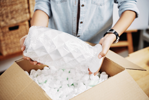 Hands of woman putting fragile vase in box filled with foam pettels