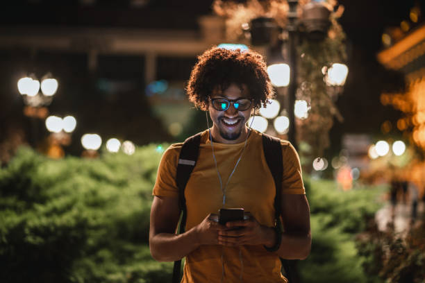 Smiling African student texting outside Young African student is texting message on smart phone outdoors at night. mobile phone lights up stock pictures, royalty-free photos & images