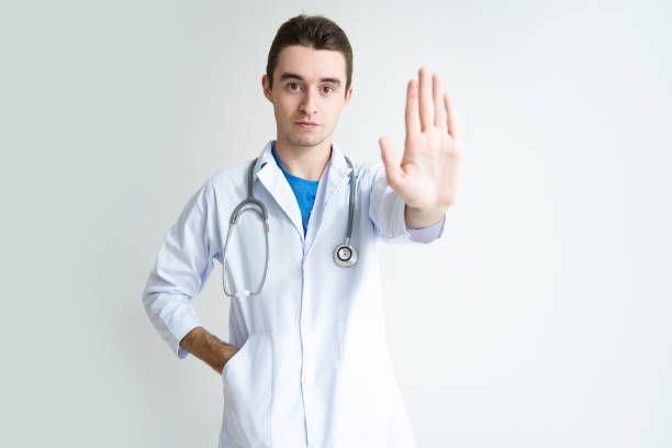 Portrait of serious young male doctor showing stop gesture Portrait of serious young male doctor showing stop gesture. Caucasian general practitioner in labcoat with stethoscope showing palm. Prohibition or refusal concept male is standing and shows outstretched hand with open palm stock pictures, royalty-free photos & images