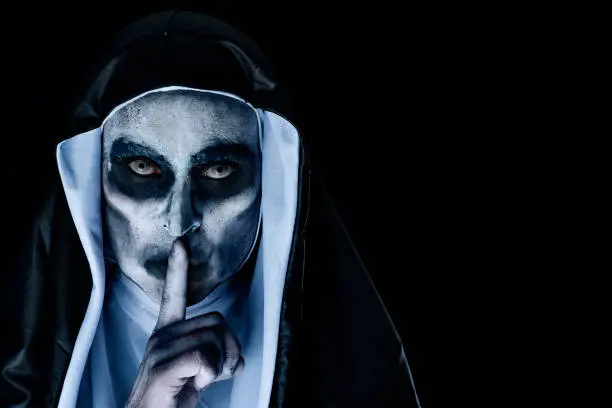 closeup of a frightening evil nun, wearing a typical black and white habit, asking for silence, against a black background with a blank space on the right