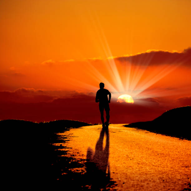 istock Silhouetted man running on dirt road over sunset sky 1049277422