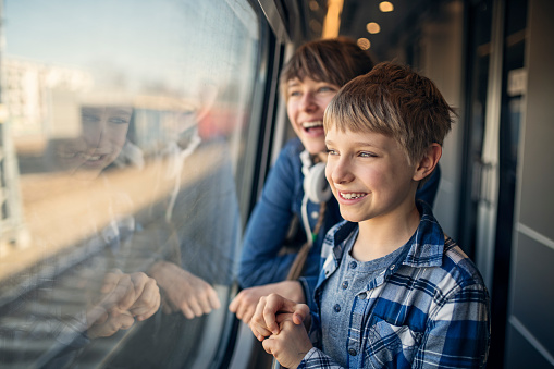 Kids travelling by train. Little boy and his teenage sister and standing by window in train corridor. Kids are talking and laughing while looking through the train window.\nNikon D850