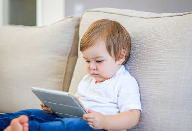 Cute little baby watching cartoon on digital tablet sitting on the sofa. Parenting control. Child internet safety concept. stock photo