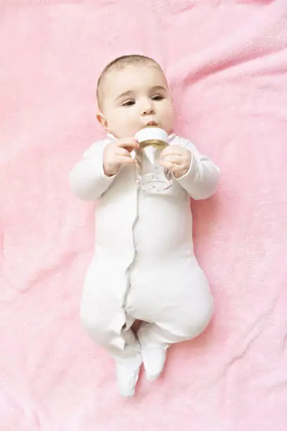 Cute caucasian baby drinking water from the bottle while laying down on the pink blanket. Vertical overhead shot