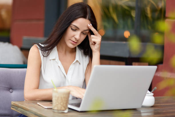 istock Serious brunette woman thinks on how make startup project correctly, wears white blouse, works with laptop computer in cafeteria, drinks milk cocktail, keeps hand on head, tries to concentrate 1049307208