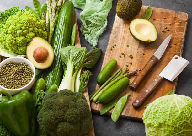 istock Assorted green toned raw organic vegetables in wooden box on dark background. Avocado, cabbage, cauliflower and cucumber with trimmed beans and chopping board with knife. 1049310824