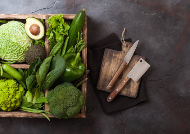 istock Assorted green toned raw organic vegetables in wooden box on dark background. Avocado, cabbage, cauliflower and cucumber with trimmed and mung beans and chopping board with knife. 1049313854