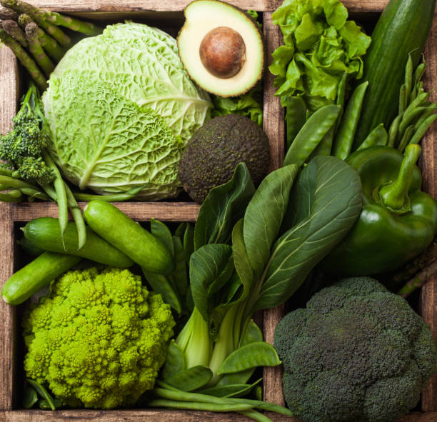 istock Assorted green toned raw organic vegetables in wooden box on dark background. Avocado, cabbage, cauliflower and cucumber with trimmed and mung beans and pepper and broccoli with sparagus tips. 1049313948