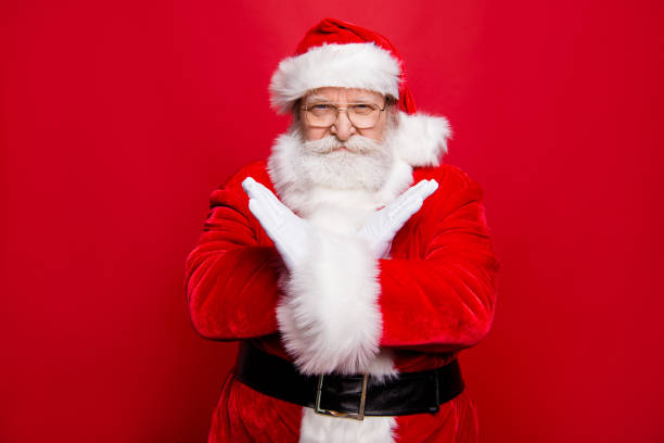 istock Concentrated confident aged stylish Santa with serious face white beard showing making gesture with crossed X hands isolated on vivid noel red background 1049346438