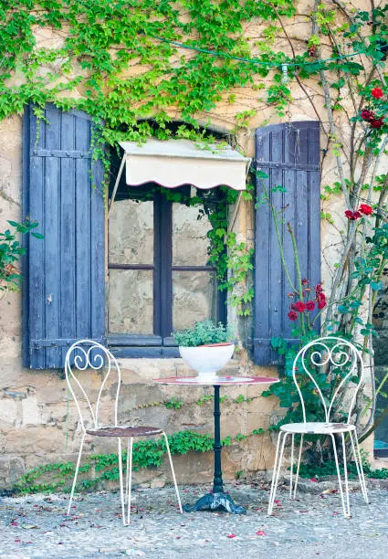 Ancient window and shutters with chairs and table outside in this narrow road by an old residence, typical architecture of the homes and houses found in the backstreet alleys between buildings of stone construction in the ancient medieval old town of Monpazier, in the department of Dordogne, southwest France