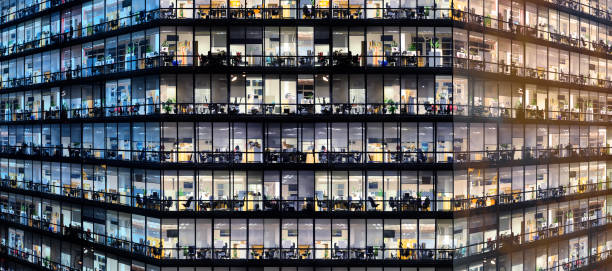 Sunset and dark sky reflecting in windows. Office workers working late in major office building The image depicts workers working late in an office at night. The employees are seen as silhouettes against their lit offices that have large windows. The building is lit by the 'blue hour' evening sky in one end and colorful cloud in the other. The building and its windows cover the entire canvas of the image. It is apparent that the building may have some sort of angle to the architecture. While most of the building emits internal light, windows also reflects the colorful sky. There is no border around the image. The windows on the edges and corners of the image are truncated in the view. office window night stock pictures, royalty-free photos & images