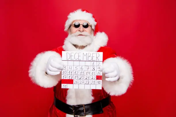 Photo of Prepare chill December noel eve. Handsome trendy careless carefree aged mature grandfather Saint Nicholas in spectacles gloves white beard hold calendar in hands isolated red background