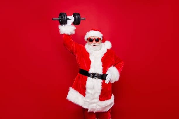 gai drôle branché élégant à la mode fort sportif viril musculaire santa à lunettes gants fourrure blanche rouge d’hiver manteau ceinture noire soulever un haltère gros s’efforçant isolé sur fond rouge - holiday healthy lifestyle weight christmas photos et images de collection