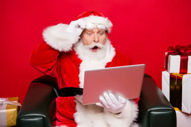 Photo of Winter noel december eve christmastime package. Aged mature Santa spectacles open mouth eyes white beard isolated red background look pc wondered astonishment face receive unbelievable email letter