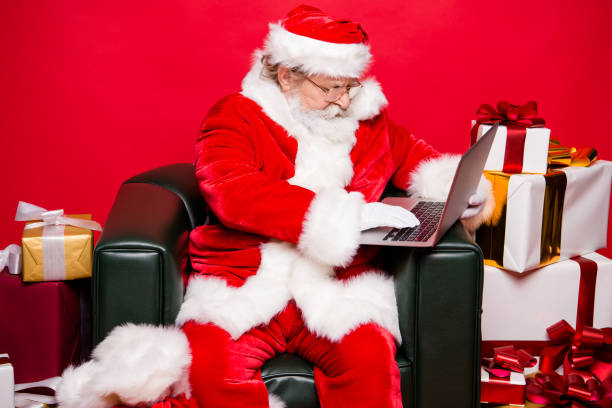 istock Christmastime winter december eve noel. Profile side view aged focused concentrated reader grandfather Santa white beard sit in armchair look at screen wish send receive email isolated red background 1049369862