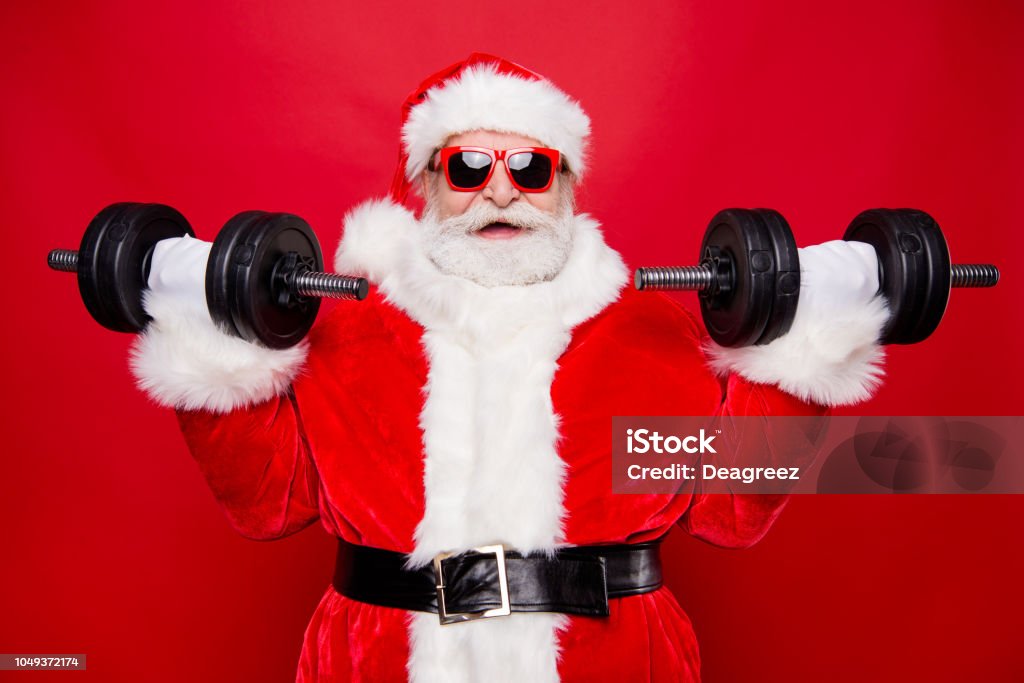 Portrait of nice optimistic positive modern stylish strong sporty muscular virile Saint Nicholas in gloves fur white red winter clothes holding lifting two big dumbbells isolated over red background Santa Claus Stock Photo