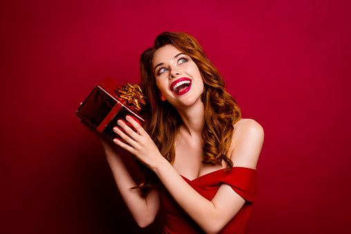 Portrait of pretty, charming, sweet, gorgeous, good-looking, alluring woman holding present box near ear, trying to know what inside isolated on dark red background