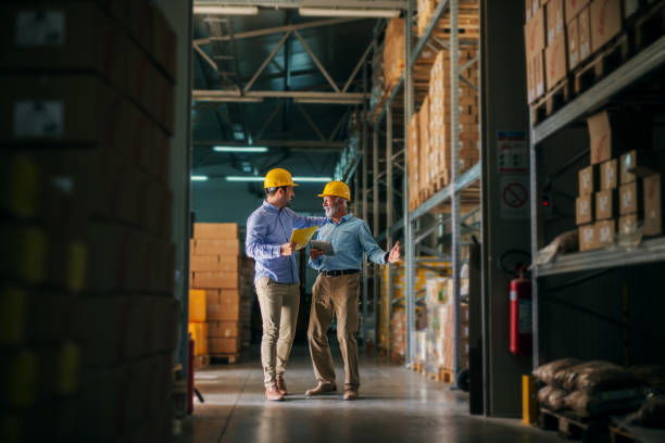 foto de dos hombre de negocios permanente en almacén con cascos en sus cabezas y celebrando buenas noticias acerca de su negocio. permanente en almacén grande y mirada feliz y satisfecho. - warehouse manager place of work portrait fotografías e imágenes de stock