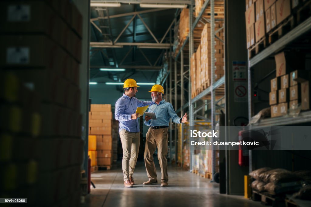 Imagens de dois homem de negócios permanente no armazém com capacetes na cabeça e comemorando grandes novidades sobre seus negócios. De pé no grande armazém e olhando feliz e satisfeito. - Foto de stock de Frete royalty-free