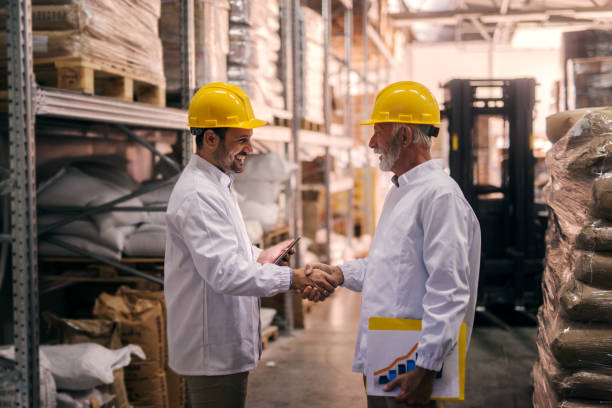 business is done. two satisfied smiling males shaking hands after closing a successful business deal. standing in big warehouse dressed in white coats with helmets on their heads. - manual worker handshake industry warehouse imagens e fotografias de stock