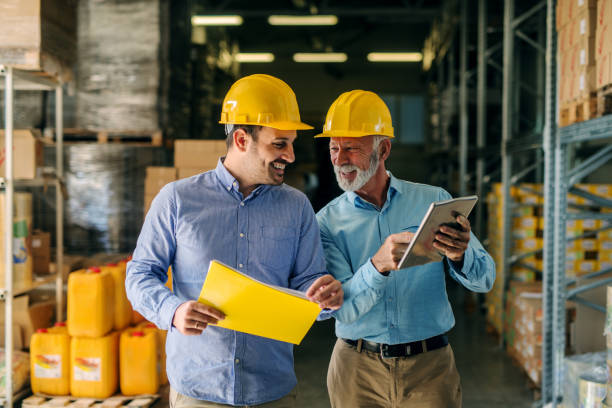 due uomini d'affari sorridenti di successo che camminano attraverso un grande magazzino con caschi in testa. l'uomo più anziano tiene in mano il tablet digitale e spinge uno più giovane alcuni documenti. - business relations foto e immagini stock