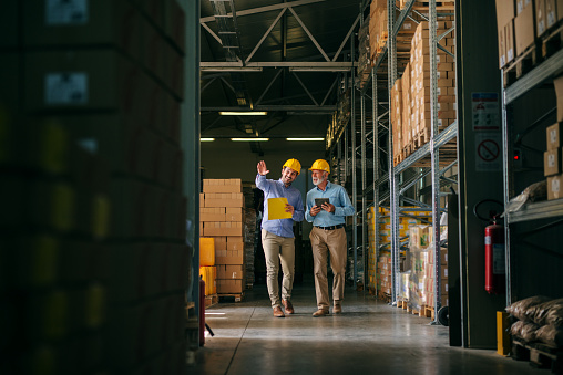 Two successful smiling business man walking through big warehouse with helmets on their heads.Younger man is shoving older one shelf’s full of products ready to be delivered. Happy investors.