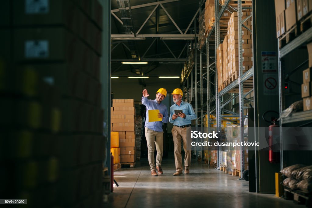 Two successful smiling business man walking through big warehouse with helmets on their heads.Younger man is shoving older one shelf’s full of products ready to be delivered. Happy investors. Warehouse Stock Photo